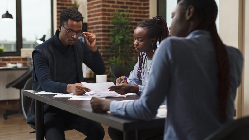 African american research agency coworkers in meeting about startup project financial status. Business people at desk in company office finance department talking about marketing expenses.
