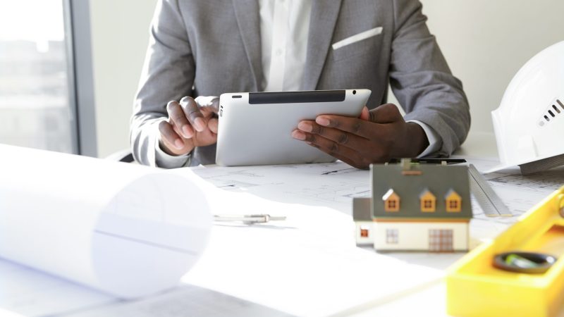 Selective focus. Design and architecture. Cropped shot of African architect holding digital tablet, developing new real estate project, sitting at desk with architectural tools and scale model house