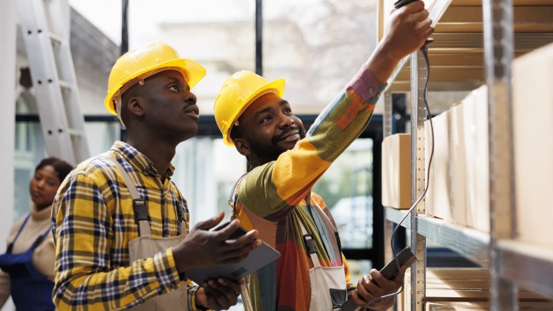 Warehouse smiling colleagues scanning cardboard box barcode and chatting. Two african american post office storehouse workers inspecting customer parcels using scanner tool and tablet